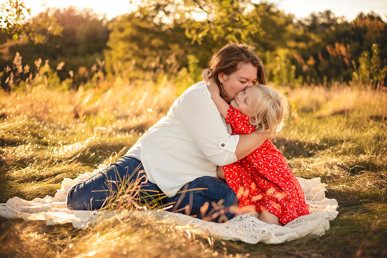 Mother kissing her daughter’s hair while hugging her on a blanket in a field during a family photography session in Bloomington-Normal