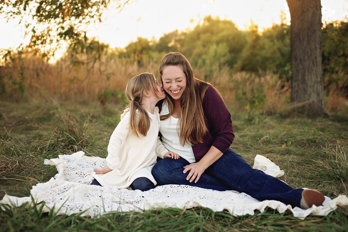 Mother and daughter sharing a secret and giggles while sitting on a blanket in a field during a family photography session in Bloomington-Normal