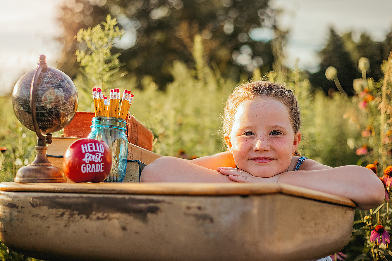 Young girl smiling beside vintage school books, a globe, pencils, and a first-grade apple during a back-to-school portrait session in Bloomington-Normal, Illinois.